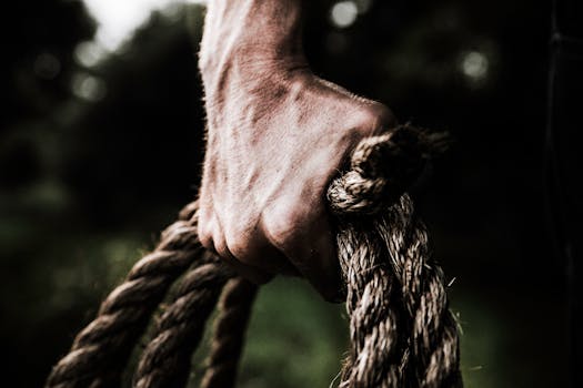 A close-up image shows a strong, firm hand gripping a thick, rough rope, showcasing strength and determination in an outdoor setting.